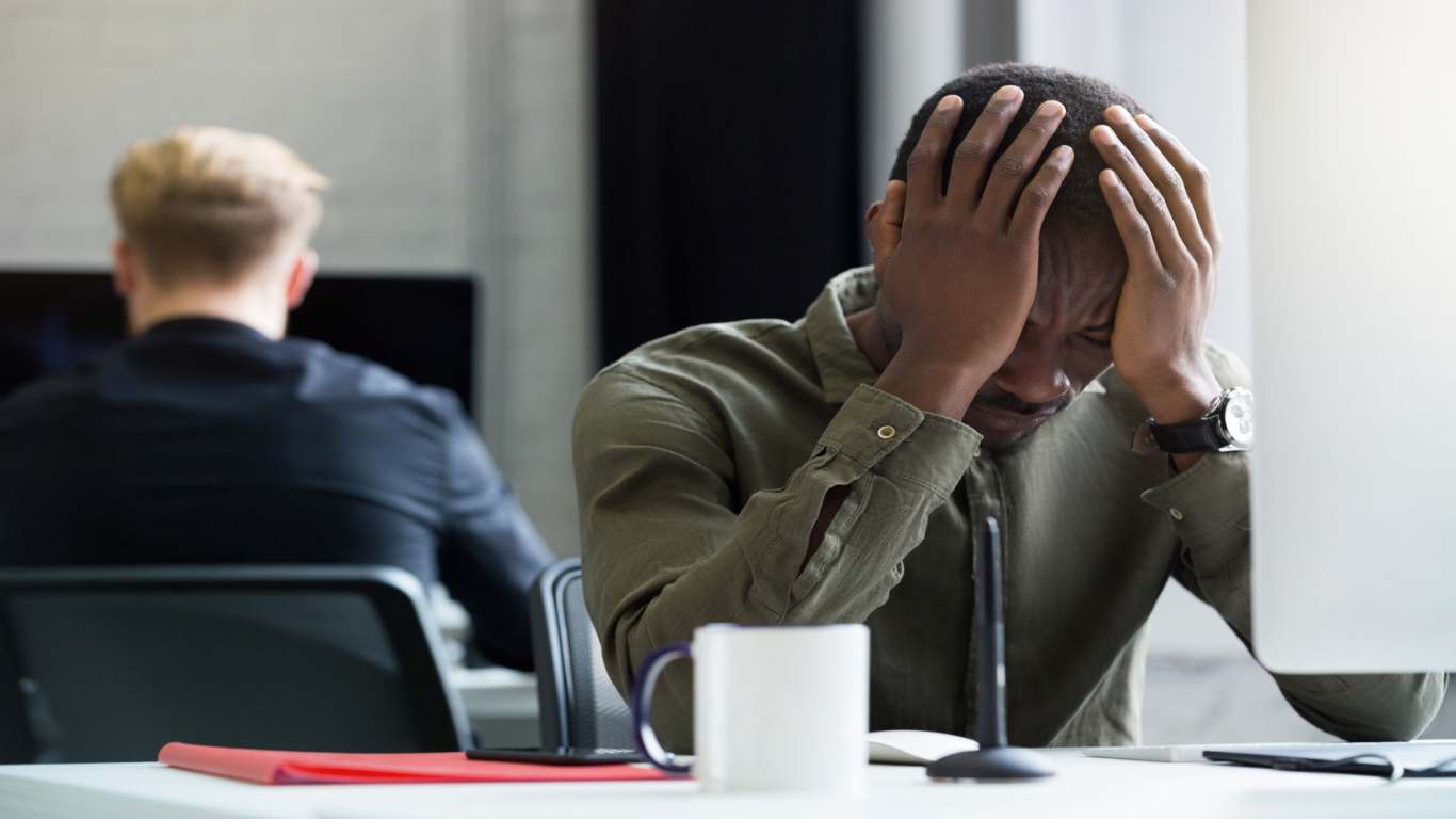 imagem de um homem sentado em uma mesa de trabalho colocando as mãos no rosto como se tivesse estressado ou chateado