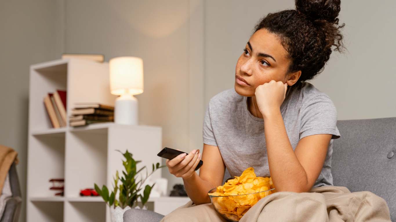 imagem de uma mulher sentada no sofá com um pote de batata olhando para a tv